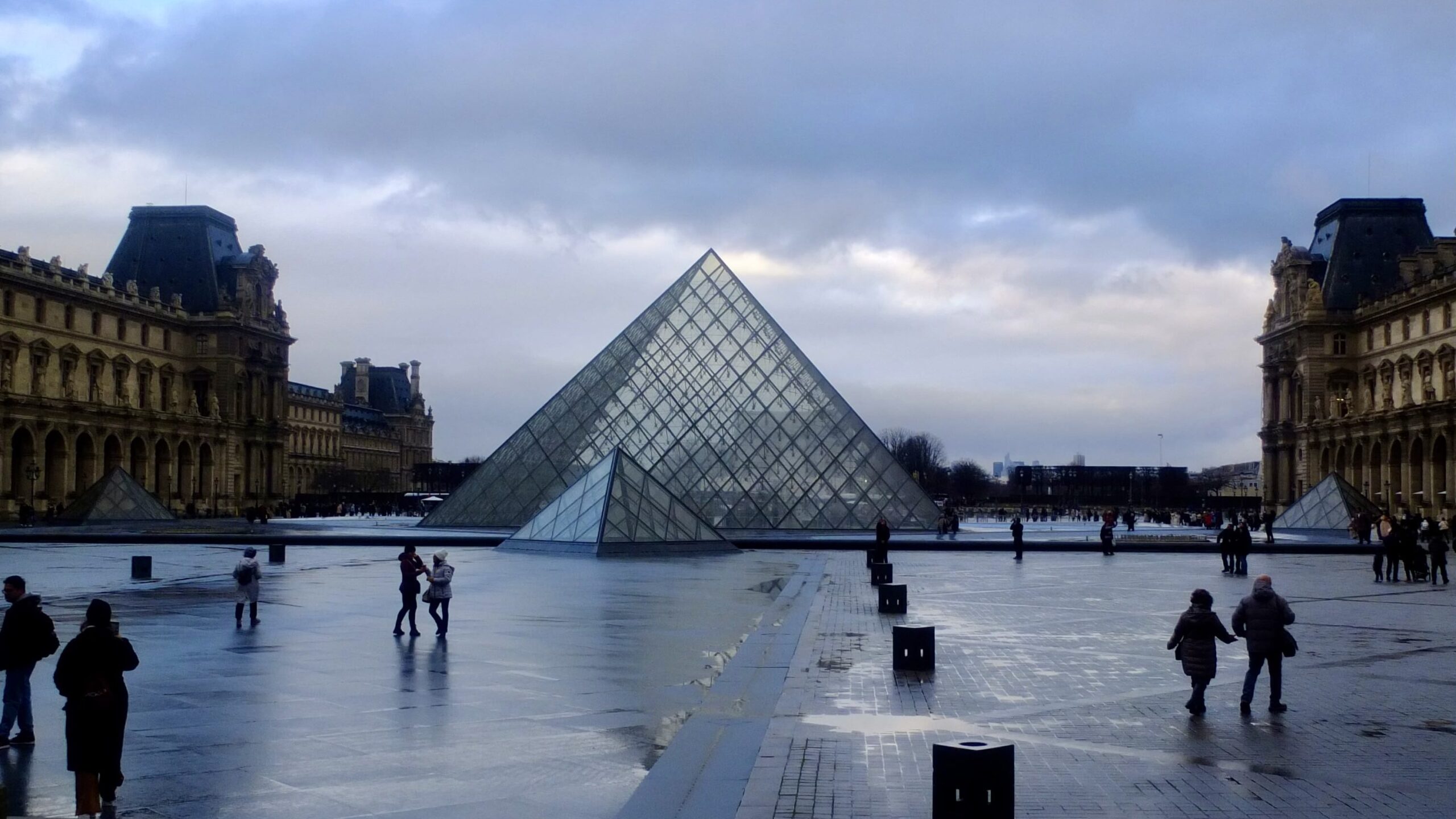 Wide view of the glass Louvre Pyramid and smaller side pyramids in a wet courtyard, framed by the palace wings.