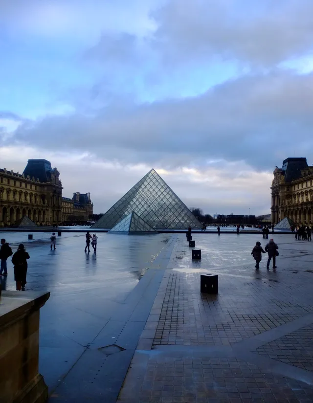 The Louvre Pyramid in a wet stone courtyard, with the palace wings on either side under an overcast sky.