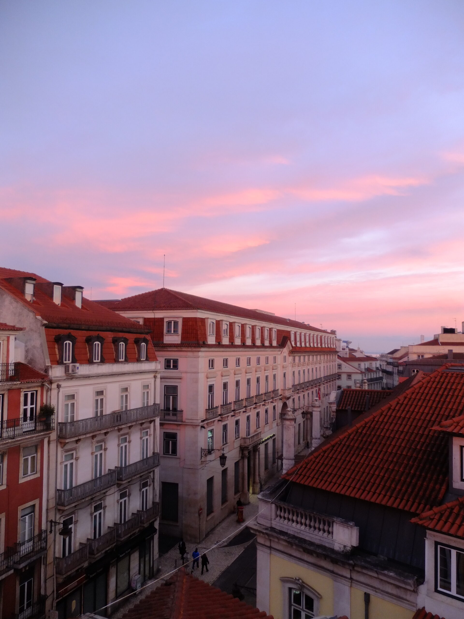 Pink twilight over terracotta rooftops and a cream neoclassical facade, with a narrow street below.