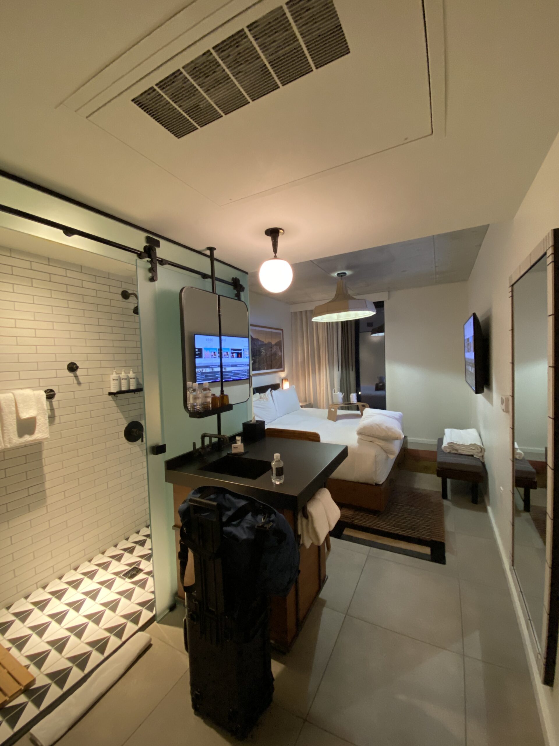 Compact hotel room with an open tiled shower, black vanity, and a bed at the far wall.