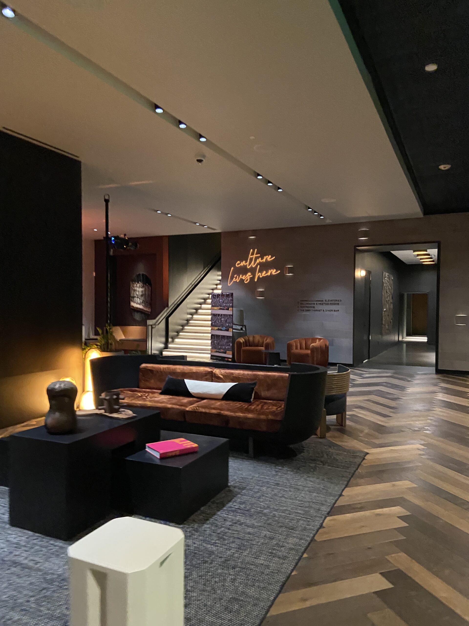 Hotel lobby lounge with a dark curved sofa, a lit staircase, and an amber neon sign reading culture lives here.