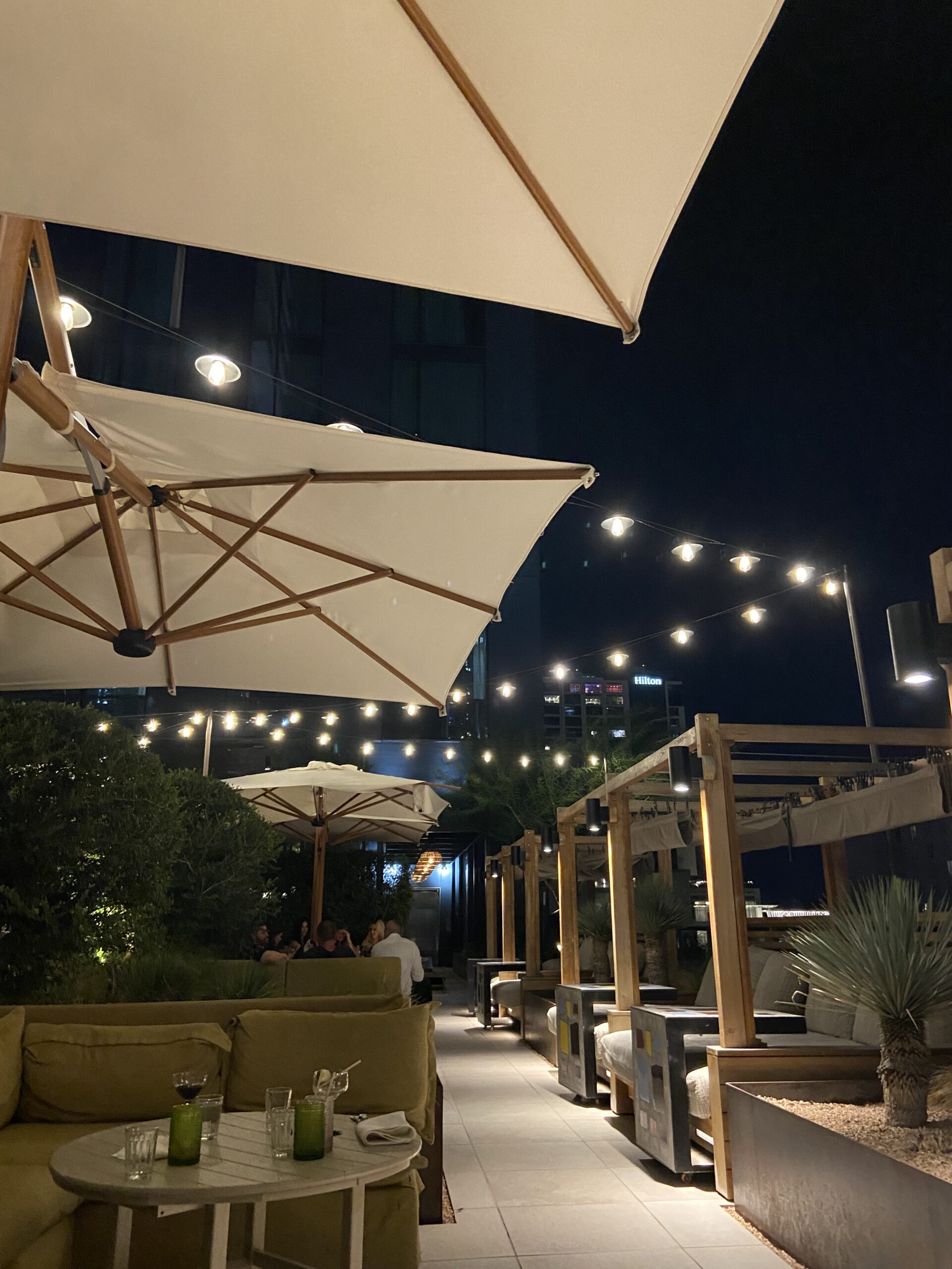 Nighttime rooftop lounge with a cream umbrella, wooden cabanas, string lights, and a city skyline.
