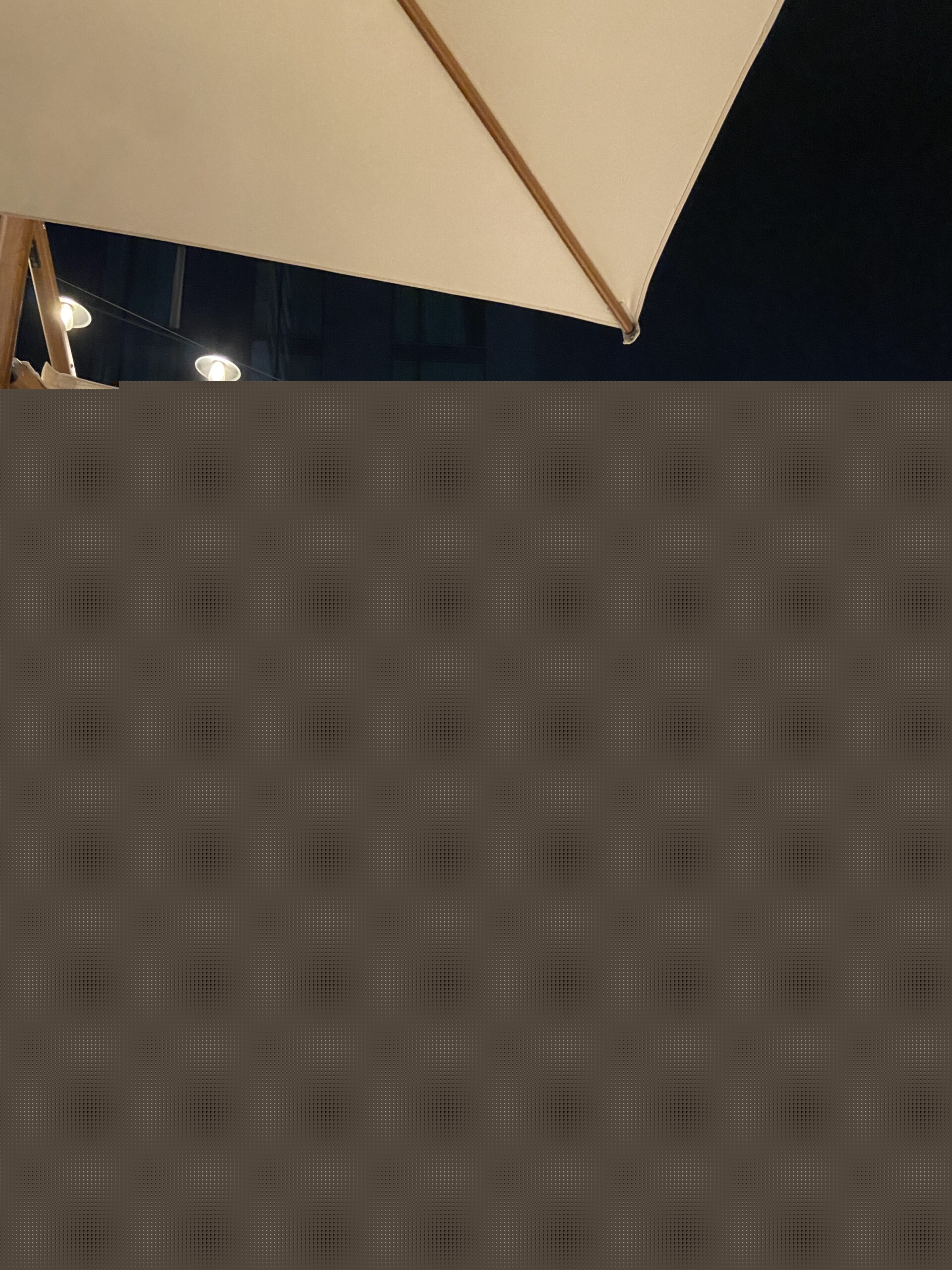 Nighttime rooftop lounge with a cream umbrella, wooden cabanas, string lights, and a city skyline.