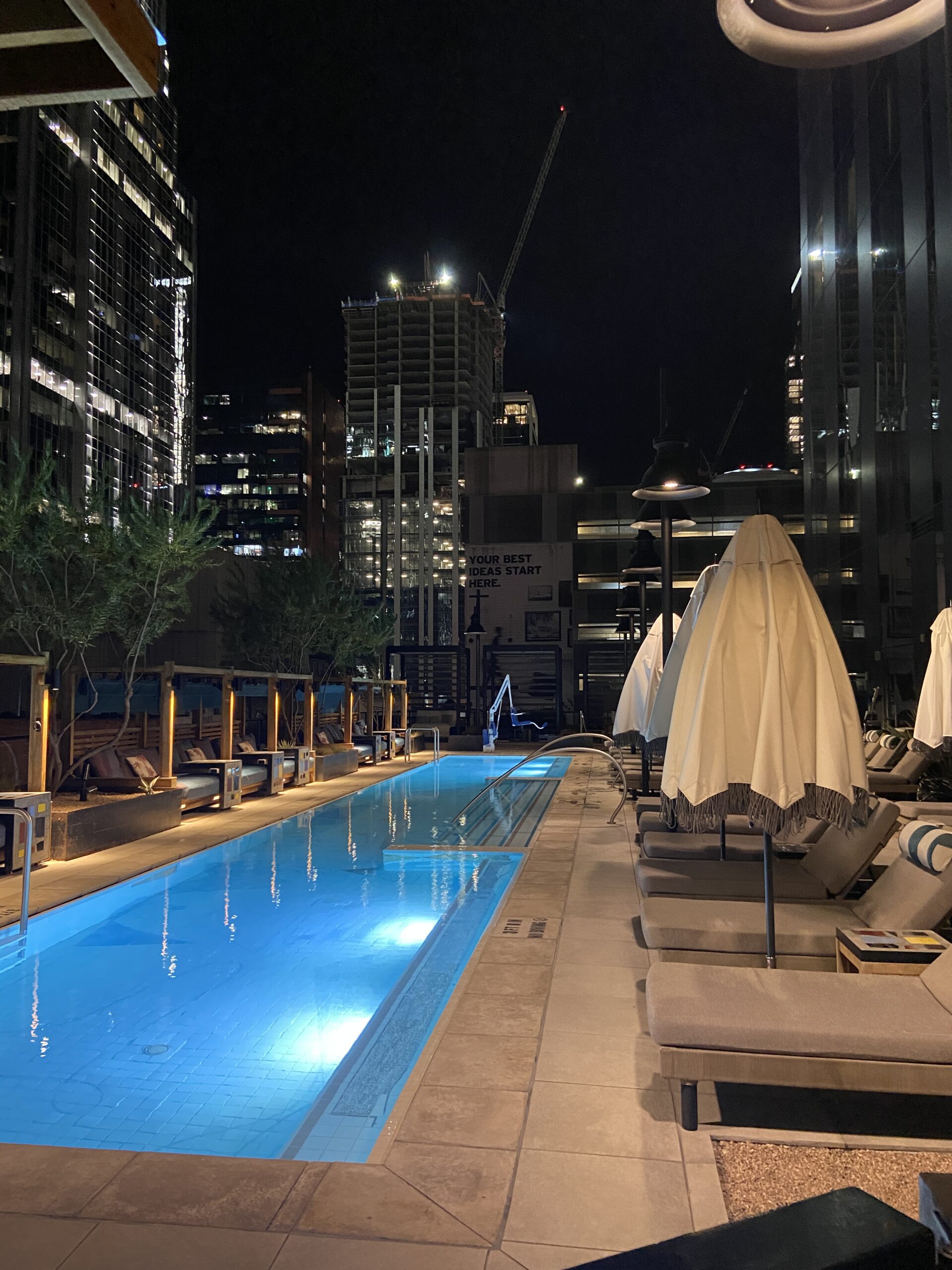 Blue-lit rooftop lap pool at night with cabanas, lounge chairs, and a construction crane on the skyline.