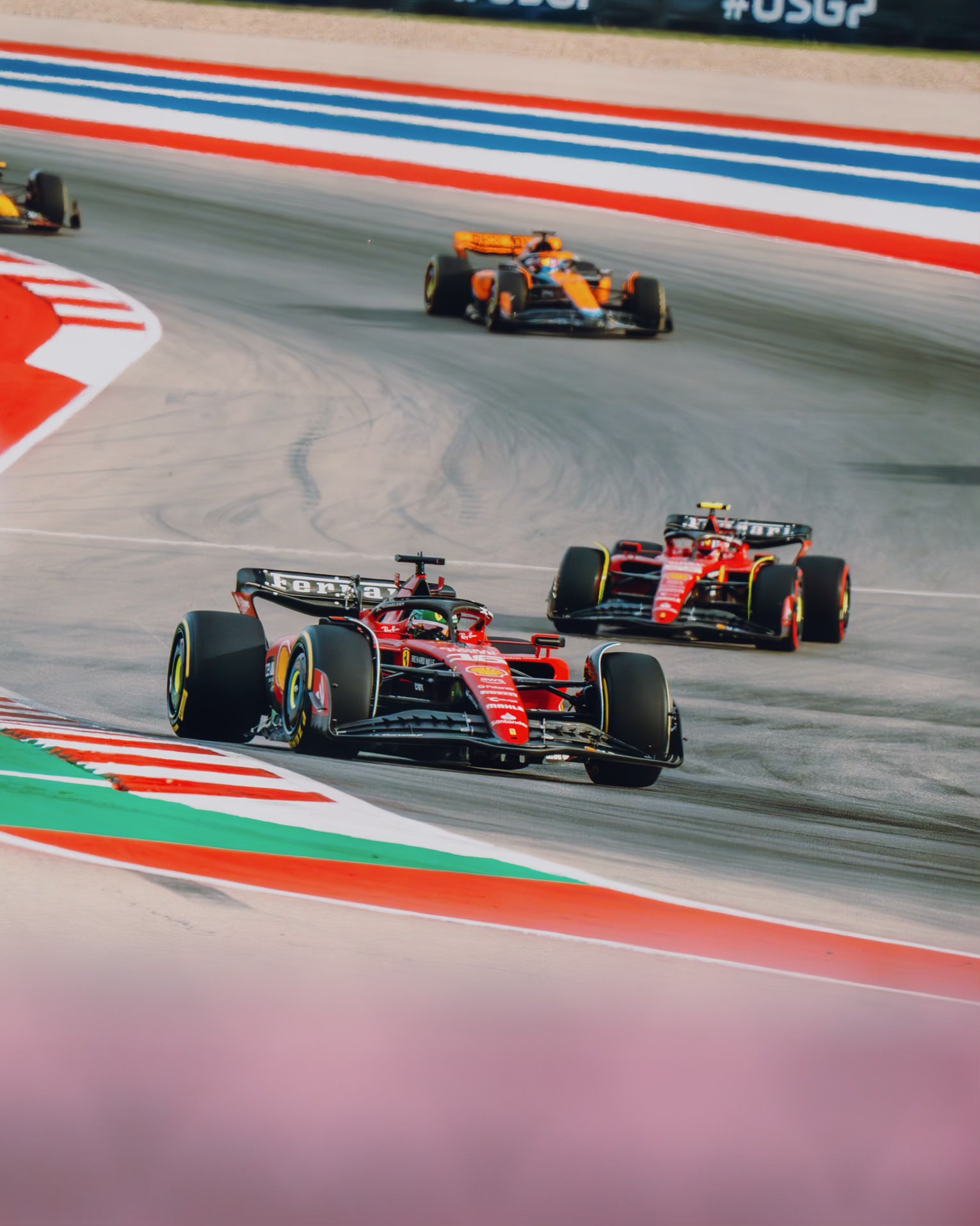 Two red Ferrari Formula 1 cars lead an orange McLaren past red-white-and-blue runoff at Circuit of the Americas.