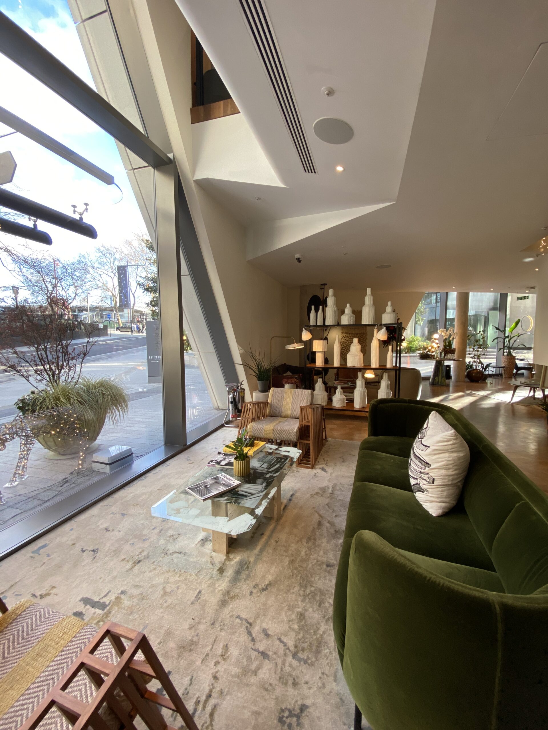 Hotel lobby seating with a green velvet sofa, slatted wood chairs, and floor-to-ceiling windows onto the street.