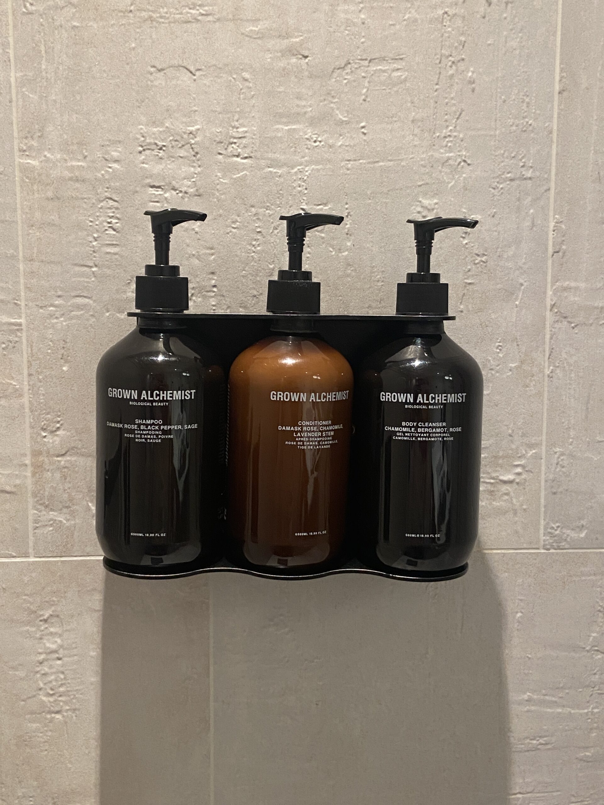 Three Grown Alchemist pump bottles—shampoo, conditioner, and body cleanser—on a black shower rack.