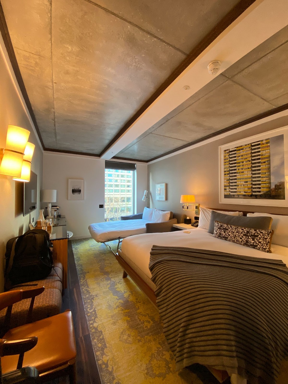 Hotel room with an exposed concrete ceiling, a made bed, and a sofa bed pulled out by the window.