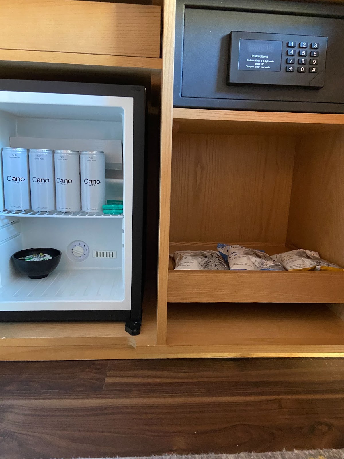 Light-wood cabinet with an open minibar of canned water, a digital safe, and snack bags in a drawer.