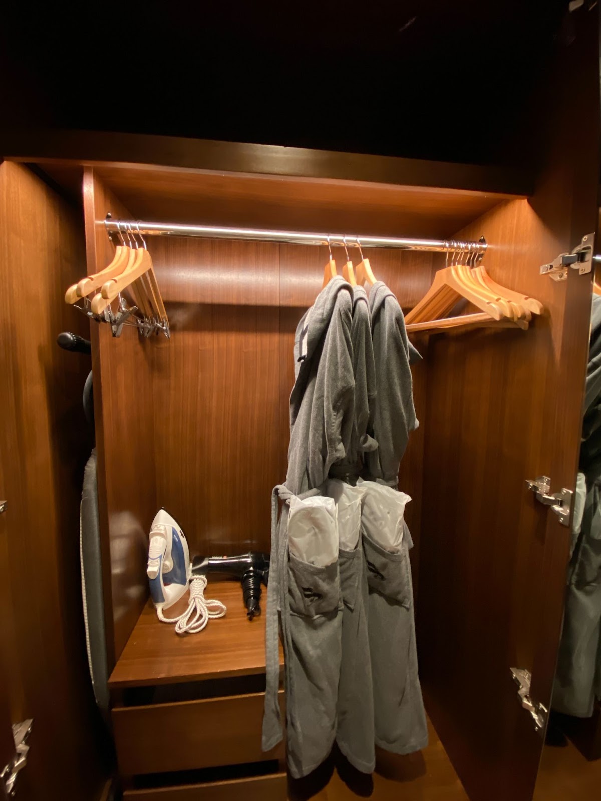 Dark wood closet with grey bathrobes, packaged slippers, wooden hangers, an iron, and a hairdryer.