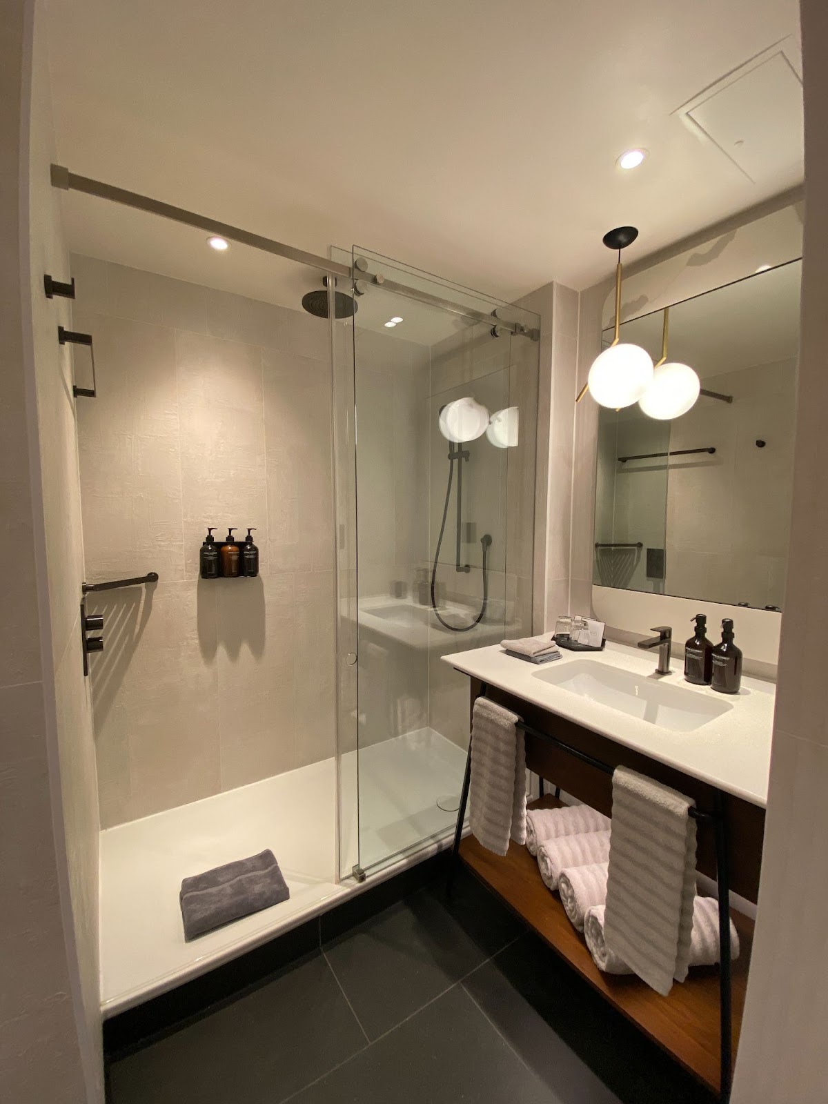 Hotel bathroom with a glass walk-in shower, amber pump bottles, and a vanity under a two-globe pendant.