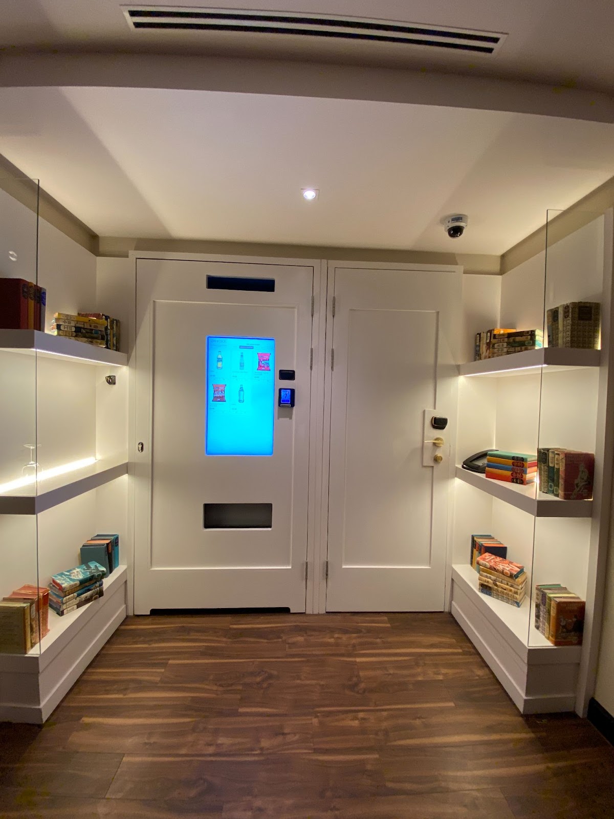 Hotel corridor with a glowing snack-vending door, a keycard room door, and backlit book shelves.