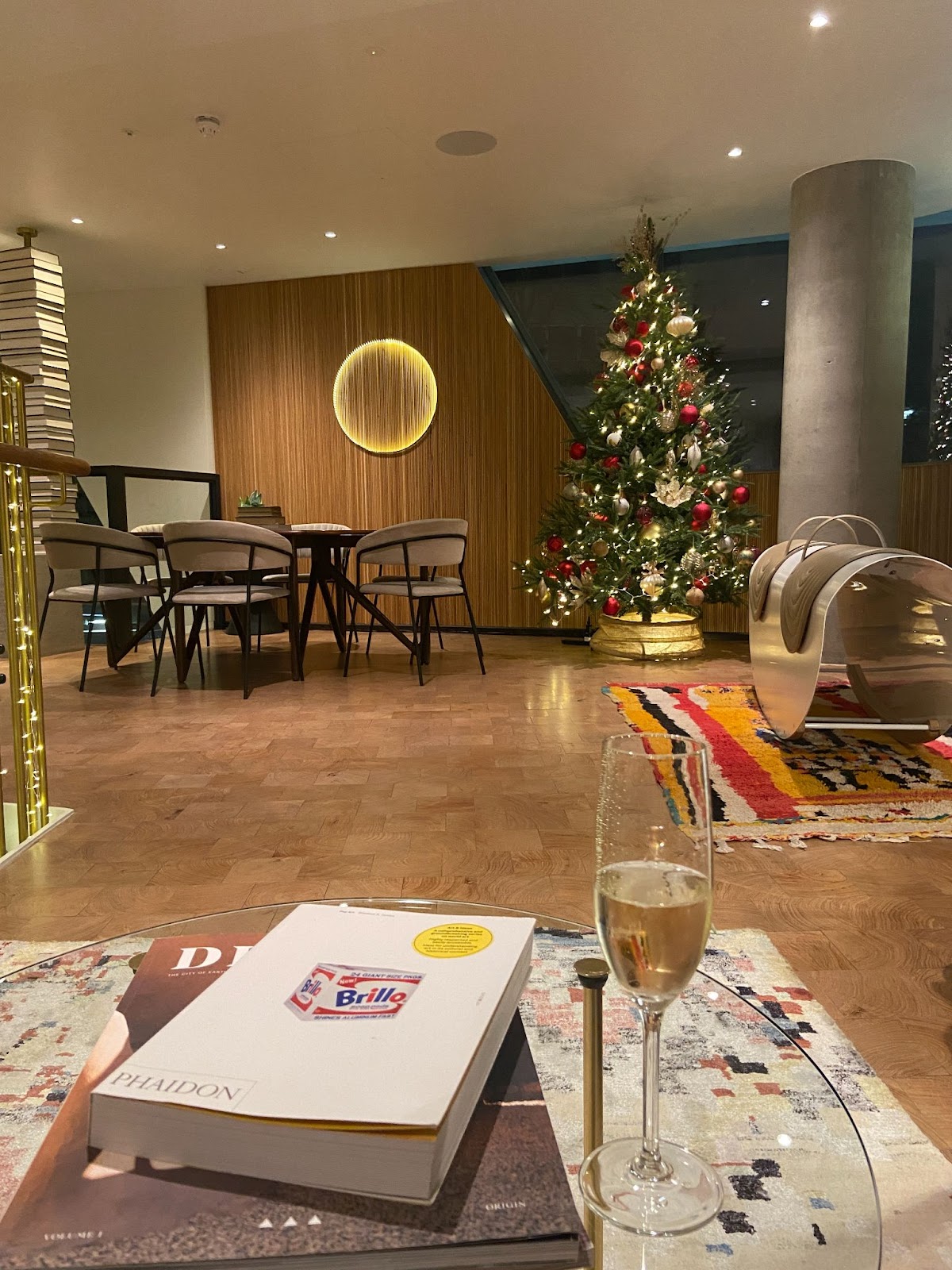 Nighttime hotel lounge with a Christmas tree, a glass of sparkling wine, and a circular glowing wall light.