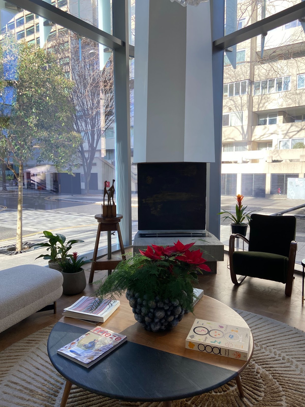 Lounge fireplace with a tall white chimney, a wooden table of magazines and poinsettias, and windows onto the street.