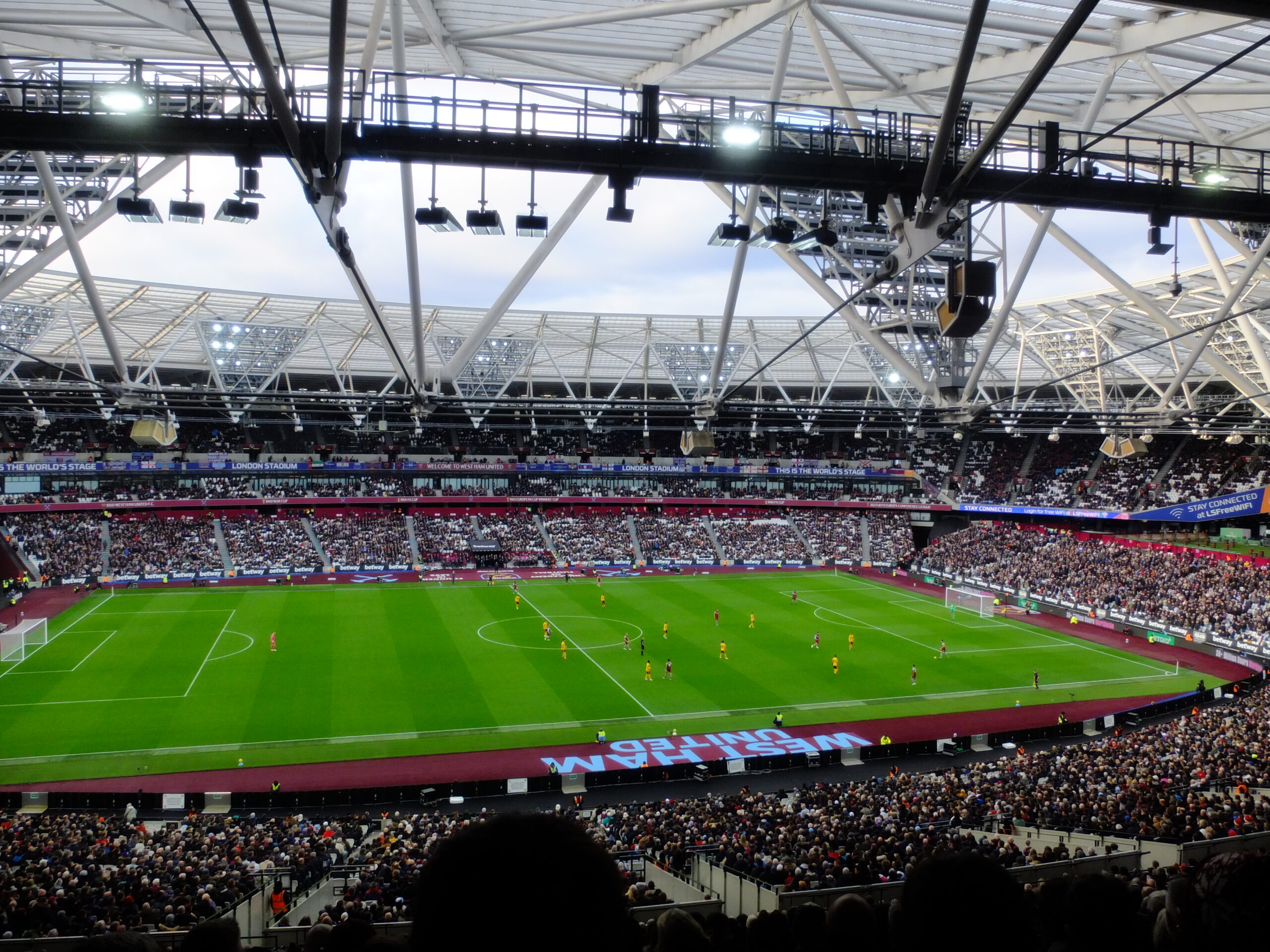 London Stadium during a match, with West Ham in claret and blue against a yellow side under the white roof.
