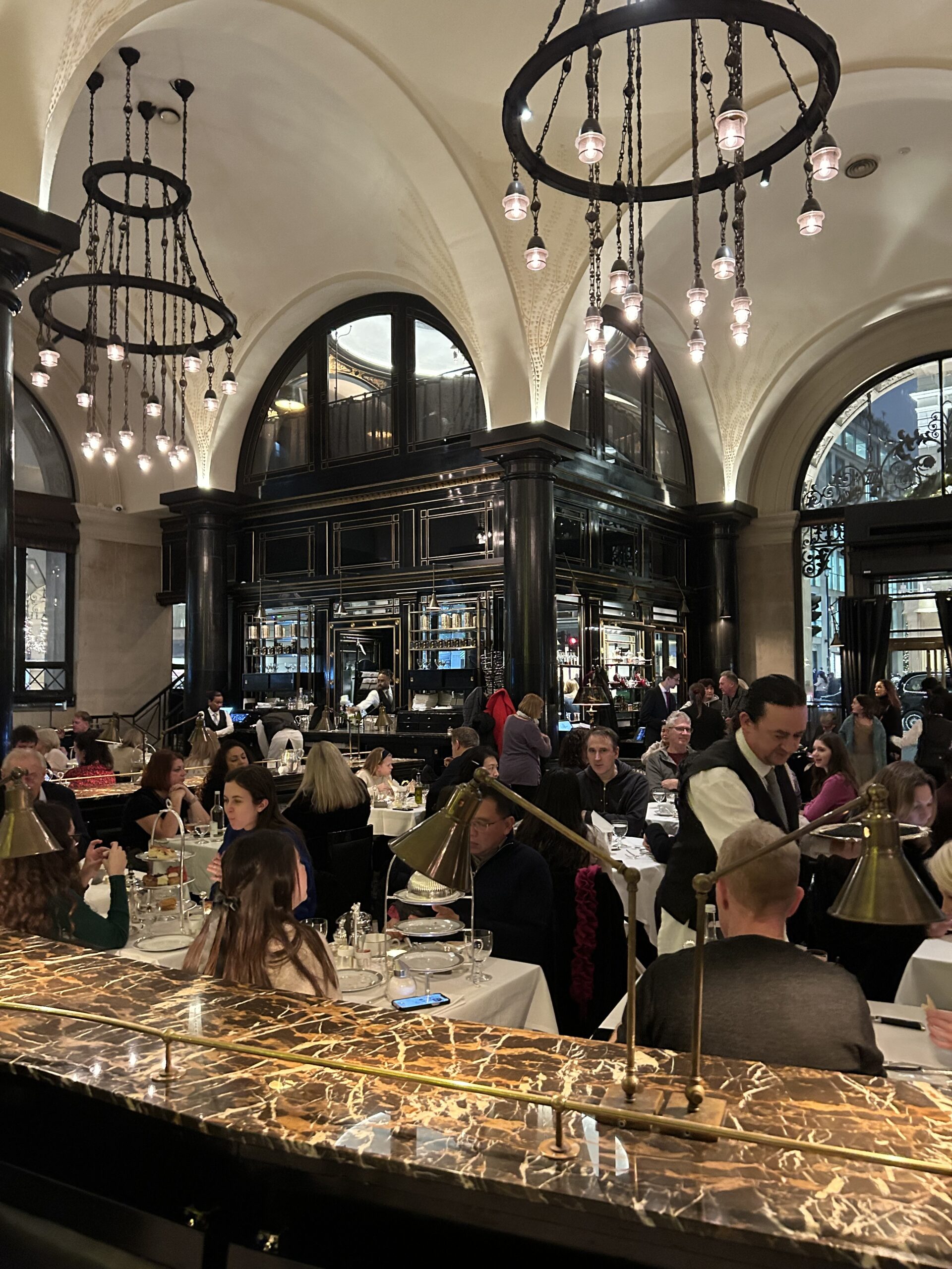 Crowded dining hall with white vaulted ceilings, black ring chandeliers, and a marble counter in the foreground.