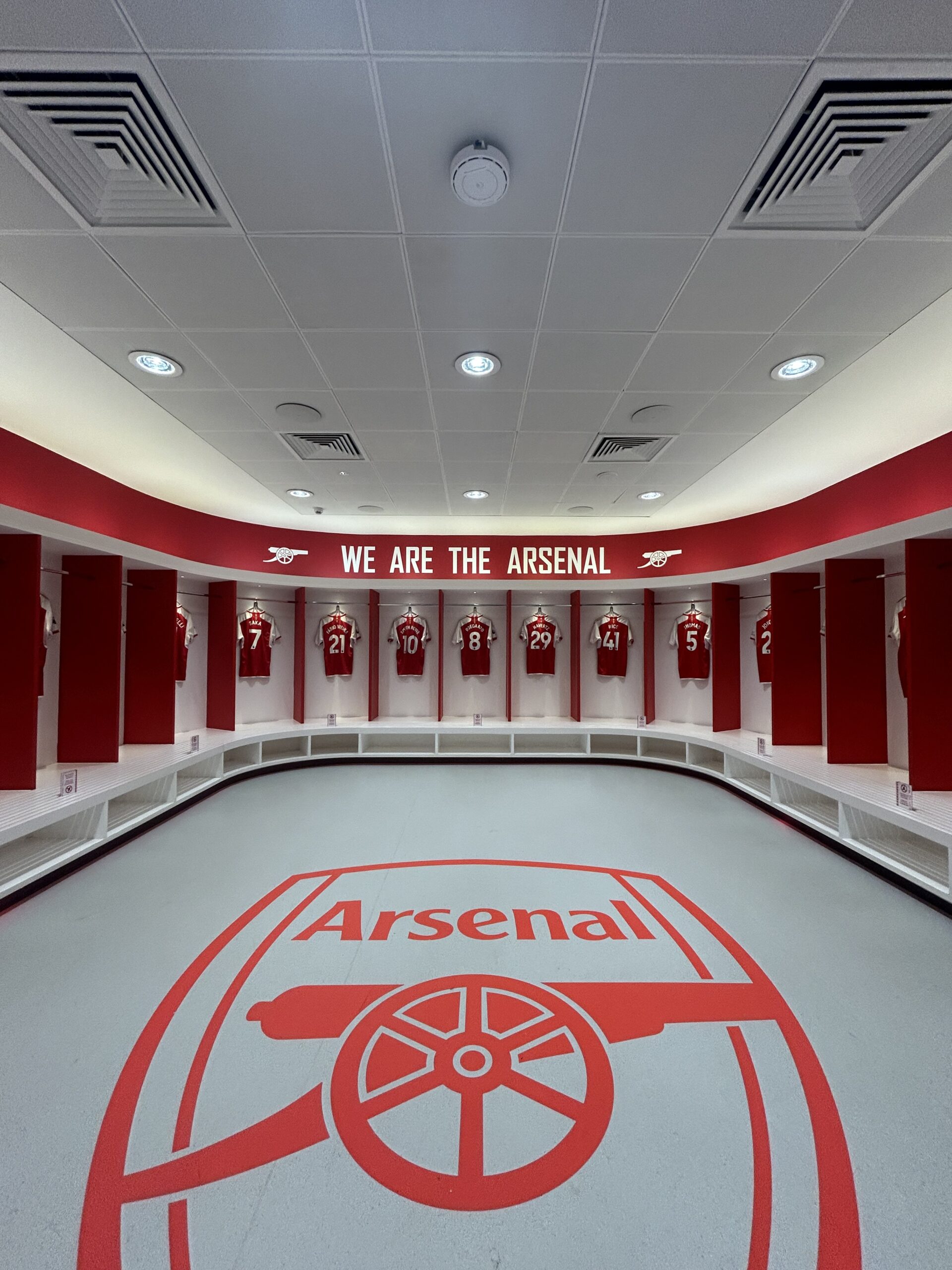 Empty Arsenal dressing room with red-and-white shirts in a semicircle of lockers and a large crest on the floor.