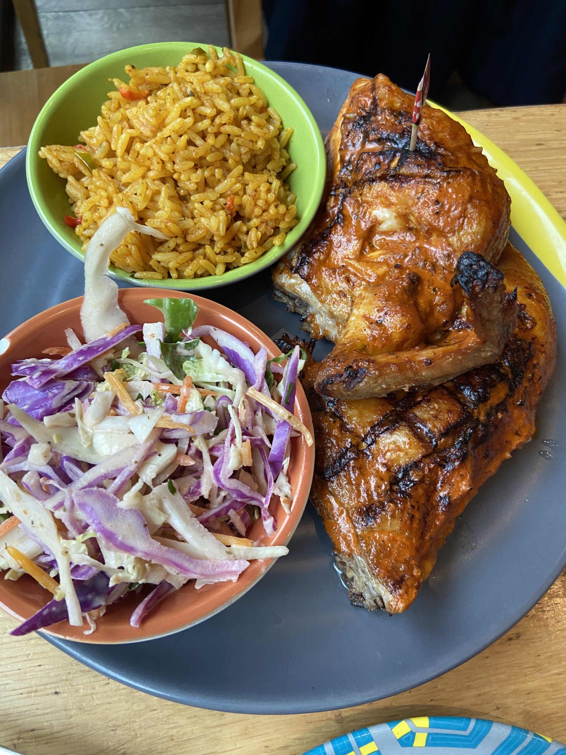 Half a charred peri-peri chicken on a dark plate with yellow rice and coleslaw in small bowls.