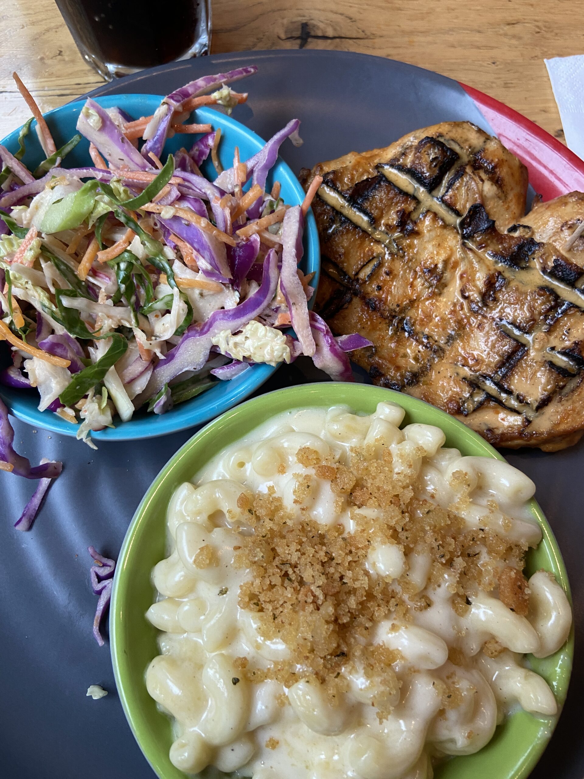 Grilled chicken breasts with a blue bowl of coleslaw and a green bowl of breadcrumb macaroni and cheese.
