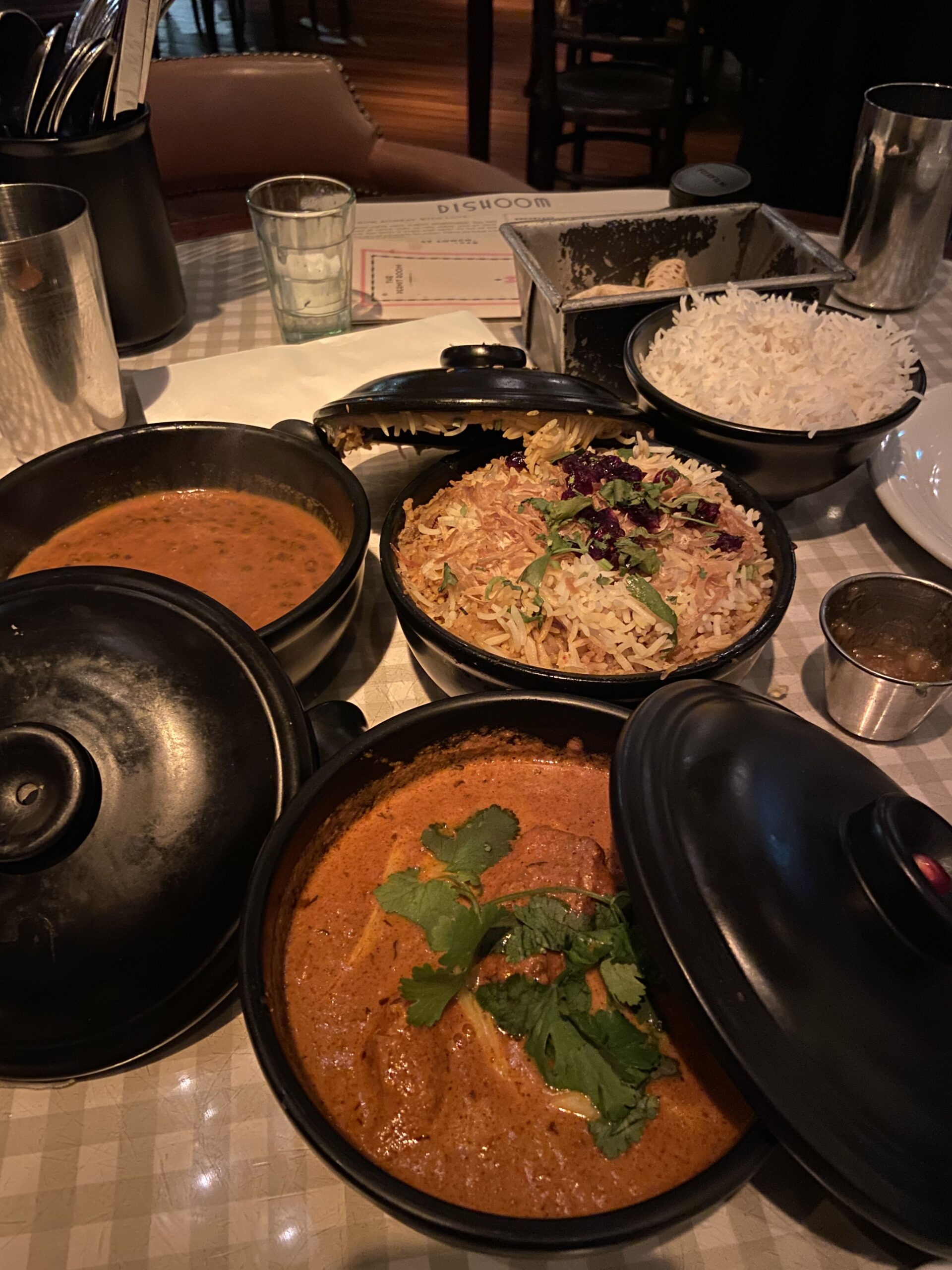 Black pots of curry, dal, and biryani on a checkered cloth beside a Dishoom menu.