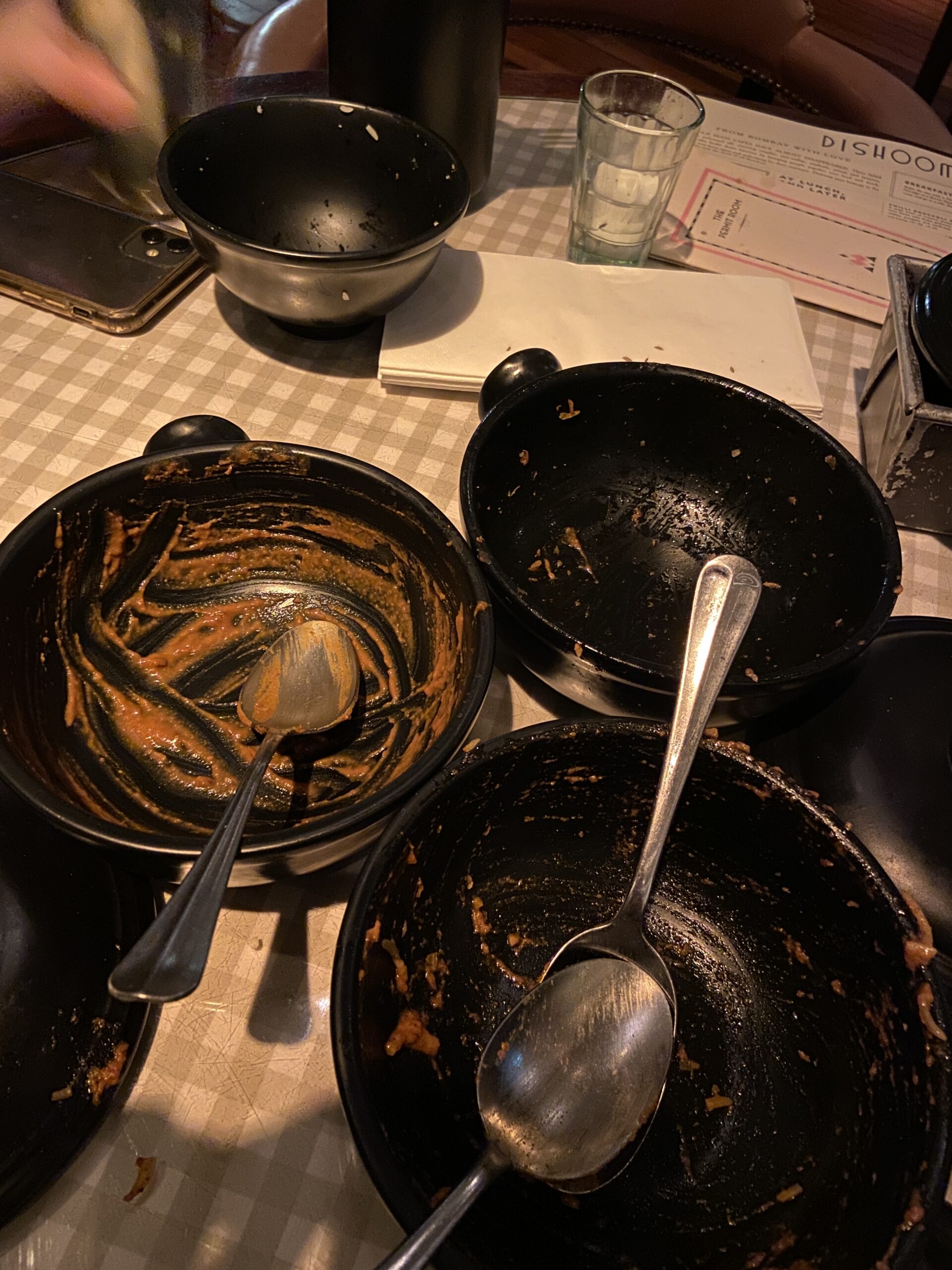 Empty black Dishoom bowls smeared with orange sauce on a tan checkered tablecloth.