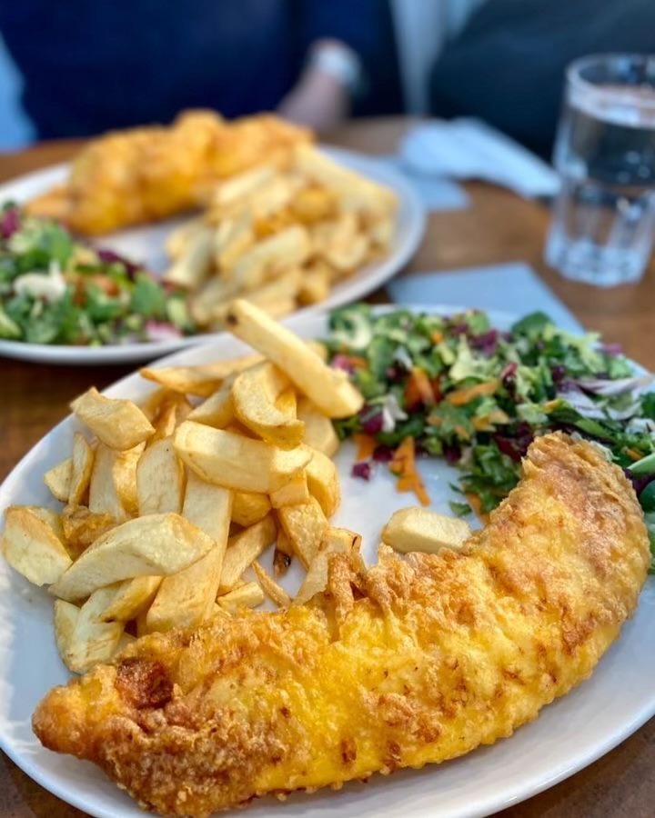 Two plates of battered fish, thick chips, and a small salad on a wooden table.