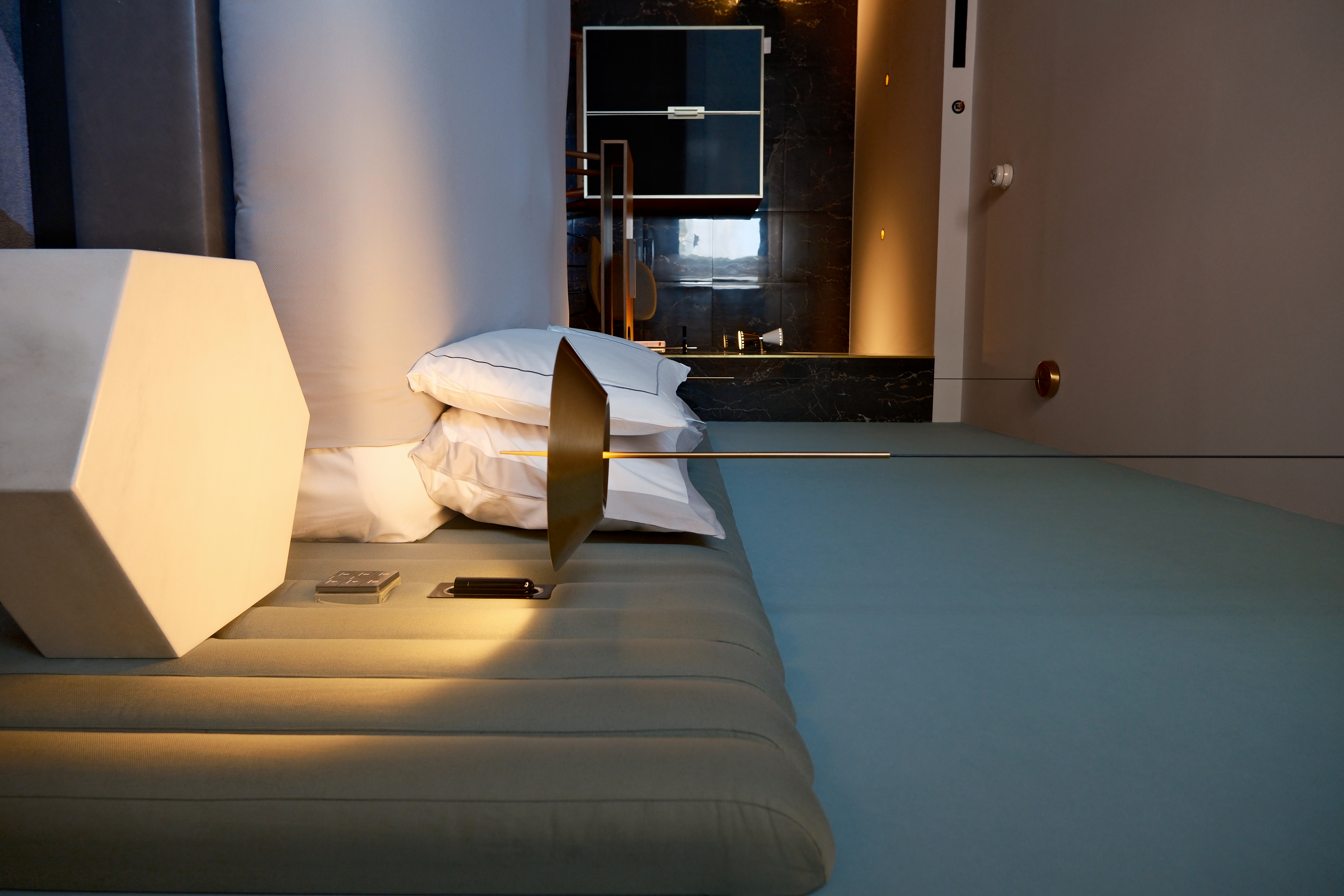 Hotel bedside with a hexagonal stone table, brass cone pendant, and a sage channel-tufted headboard.