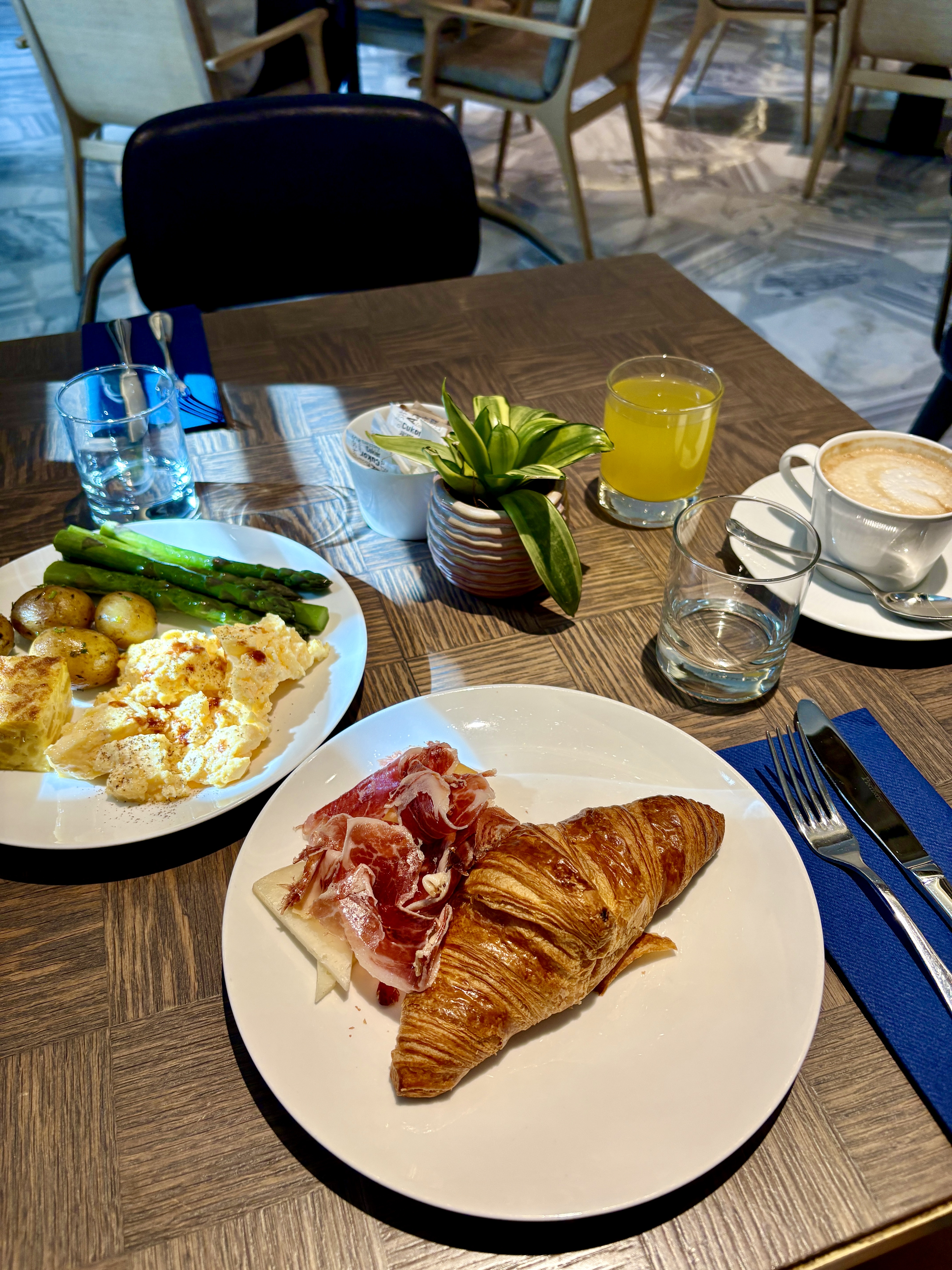 Breakfast table with a croissant and ham, scrambled eggs and asparagus, juice, and a foamy coffee.