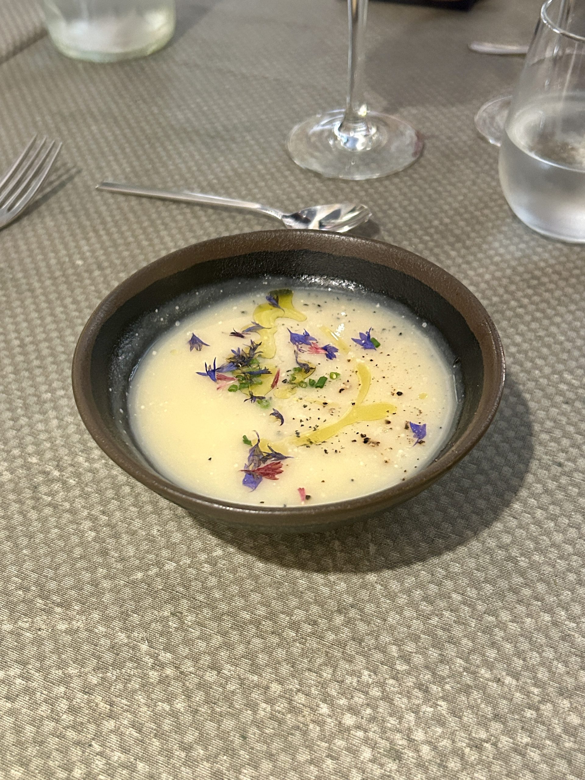 Pale creamy soup in a dark bowl, garnished with purple petals, oil, chives, and black pepper.