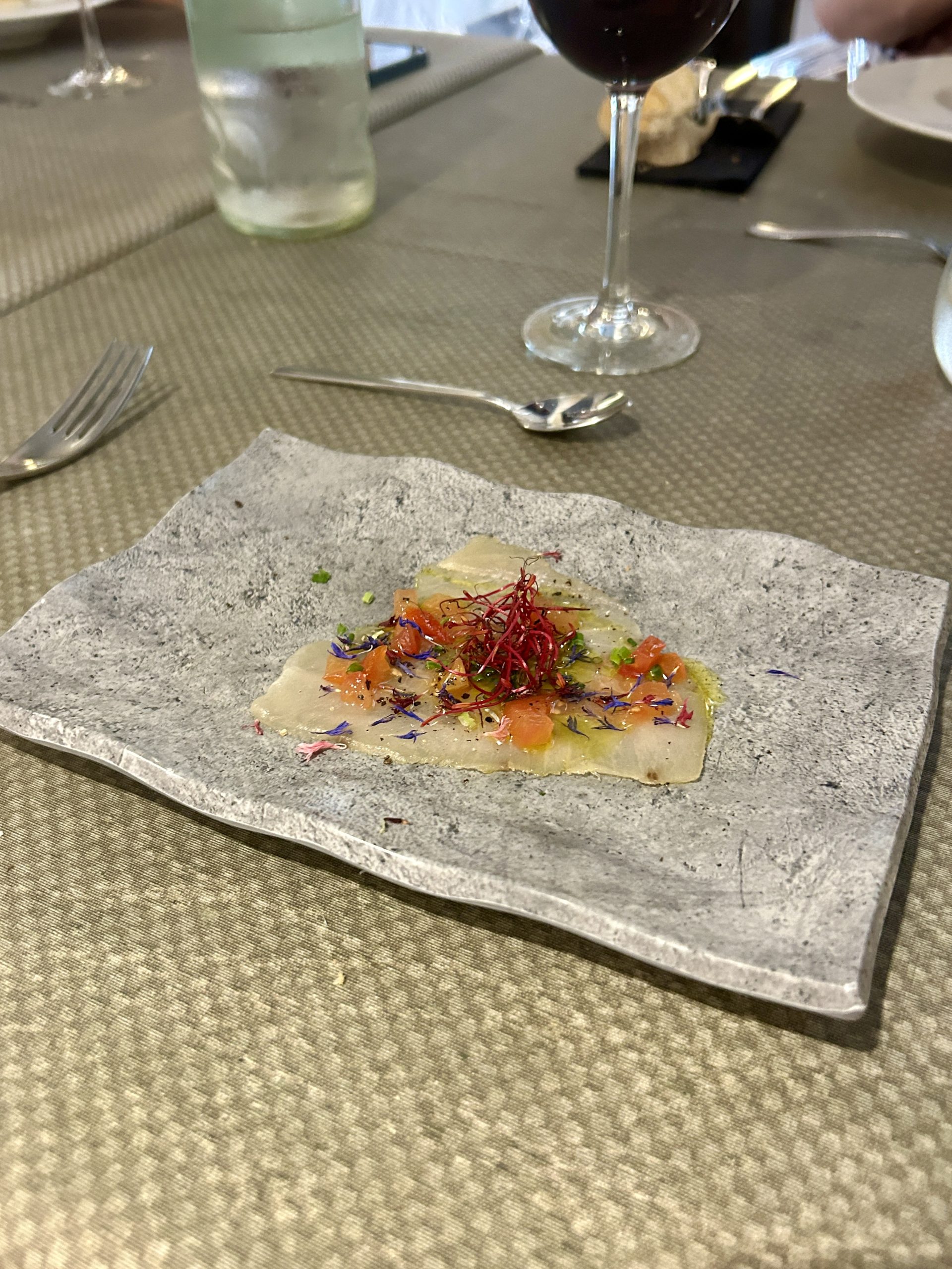 Thin slice of white fish on a grey stone plate, topped with diced orange garnish, red threads, and purple petals.