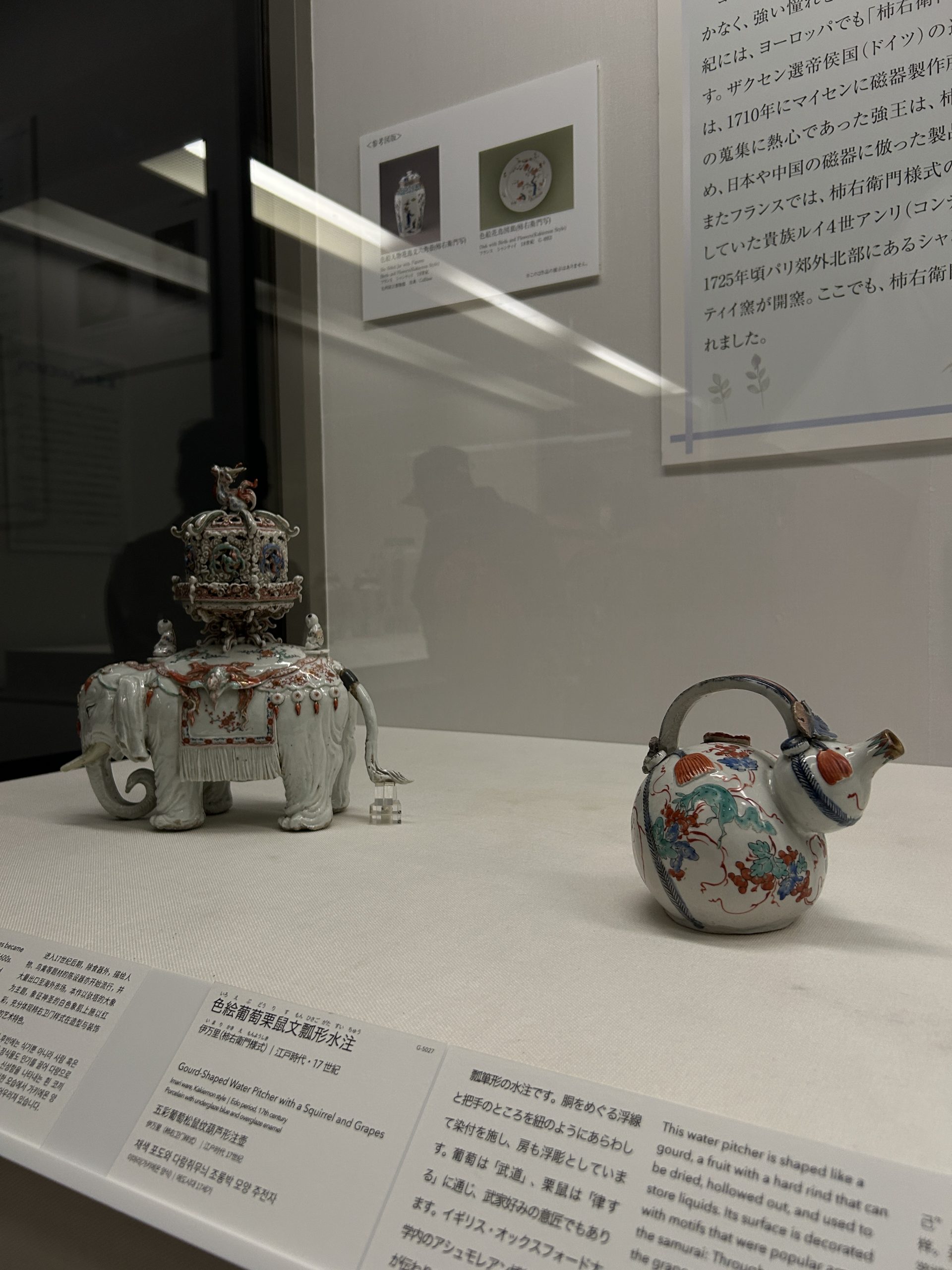 Museum case with a porcelain elephant incense burner and a gourd-shaped pitcher painted with grapes and a squirrel.