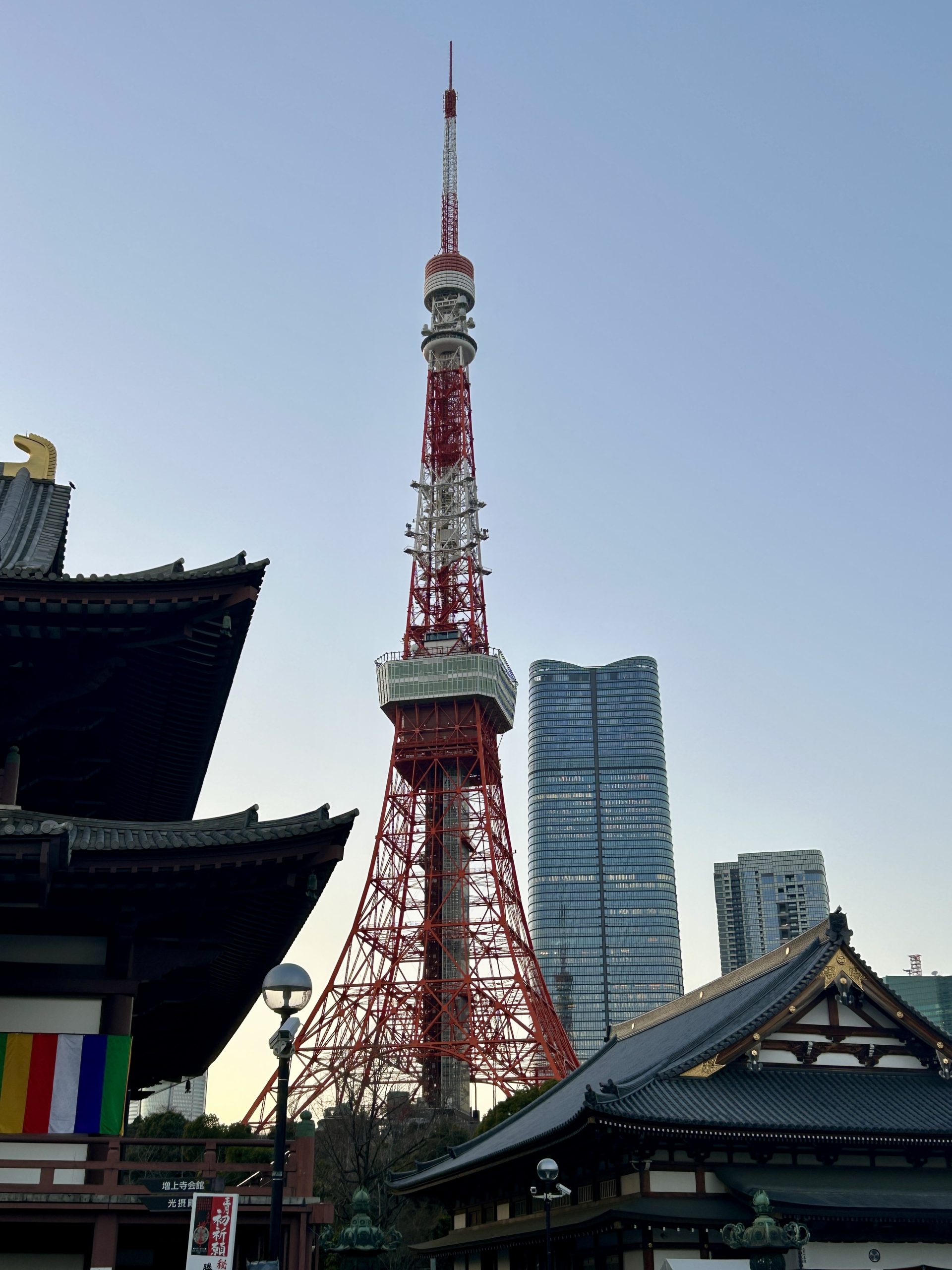 Tokyo Tower rising between temple roofs and a glass skyscraper under a clear pale-blue sky.