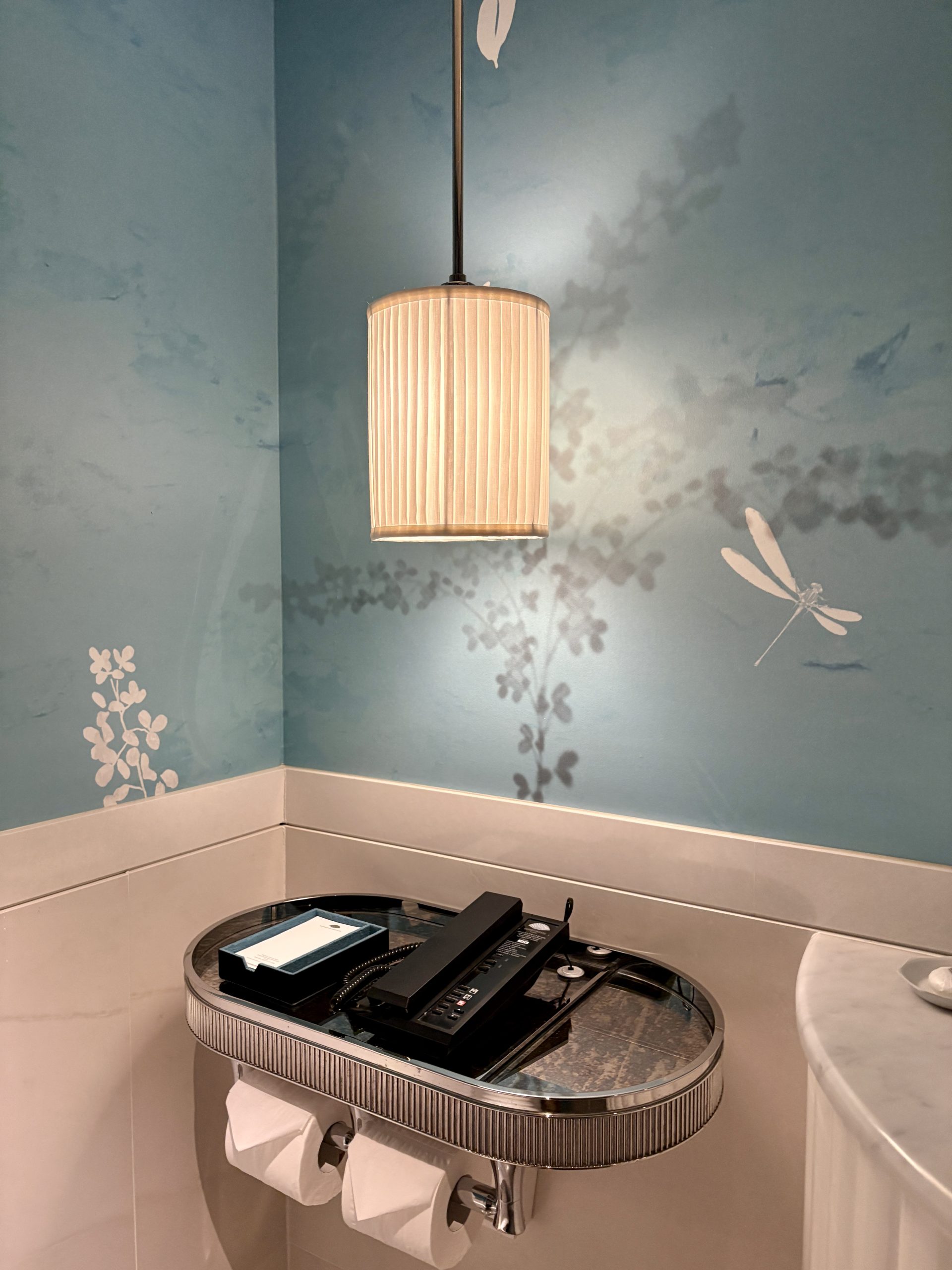 Baby blue wall paper in the wash closet with a sliver tray for phone and notepad